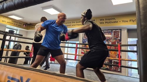 Two men sparring in a boxing ring, one in a blue shirt and the other in black, under motivational posters on the gym walls.