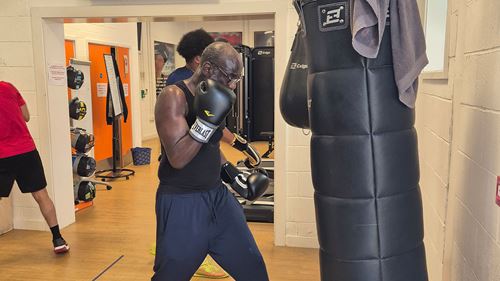 An older man wearing boxing gloves practicing punches on a standing punching bag inside a gym.