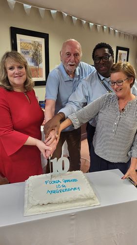 Colleagues and resident stood around cutting a white cake with 'Fiona Gardens 10yr Anniversary' written in blue on it.