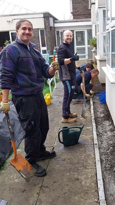 A row of people actively digging and working along a building's pathway, with shovels and a watering can.