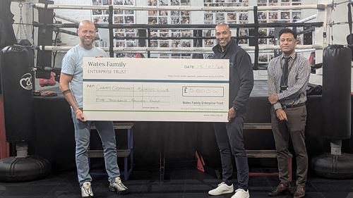 Three people in a boxing gym holding a large check for £5,000, addressed to Carney Community Boxing Club, from Wates Family Enterprise Trust.